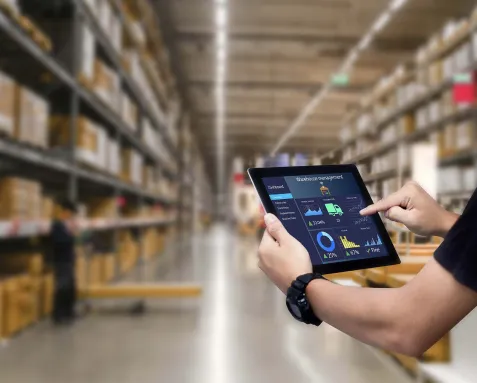 Person using a tablet to manage inventory in a large warehouse, highlighting inventory control software with features like material tracking, supply management, and budget planning.