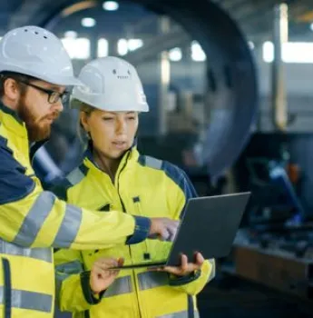 Two manufacturing workers in safety gear reviewing logistics data on a laptop in an industrial setting, representing software solutions for optimizing inventory management, production schedules, and supply chain coordination.