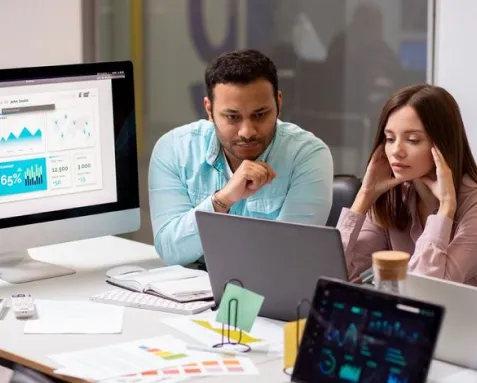 Two professionals analyzing data on a laptop and monitor, representing the development of specialized software applications like load optimization, last-mile delivery platforms, and predictive maintenance tools for logistics.