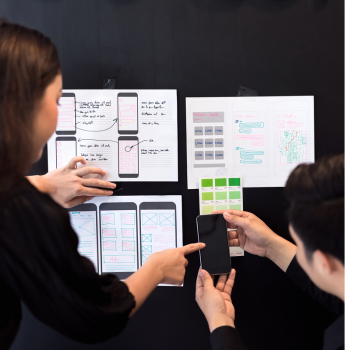 Two designers collaborating on a mobile app interface, reviewing sketches and holding a smartphone, highlighting the creation of visually appealing and engaging user interfaces using Flutter.