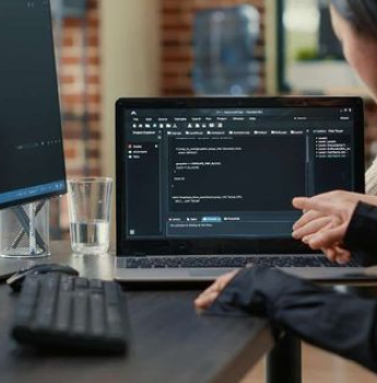 A developer pointing at a laptop screen displaying Flutter code, with an additional monitor and a glass of water on the desk in a modern office setting.