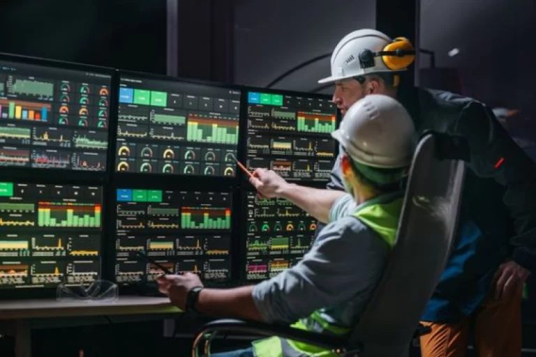 Two engineers in hard hats and safety vests monitor multiple screens displaying real-time production data in a manufacturing control room, highlighting optimized production schedules, predictive maintenance, and improved product quality.