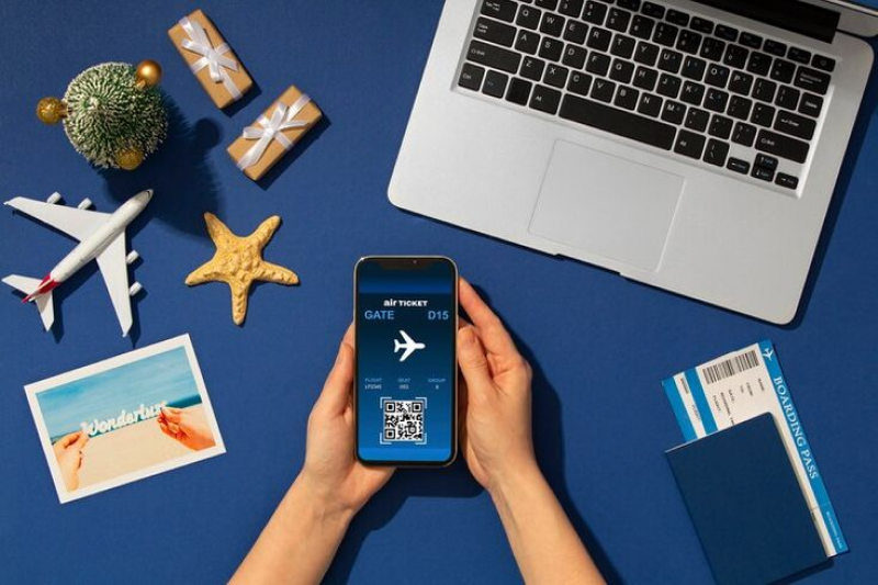 Hands holding a smartphone displaying an electronic air ticket, surrounded by travel-related items such as a laptop, boarding pass, toy airplane, starfish, and gift boxes, indicating travel planning and booking.