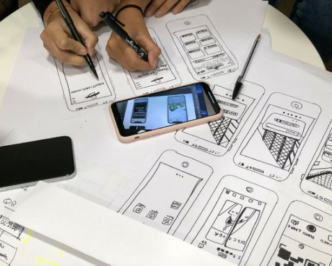 Designers sketching wireframes and prototypes for a mobile app, with a smartphone displaying an app design, highlighting the UX/UI design process including wireframing, prototyping, and iterative testing to create visually appealing and interactive user interfaces.