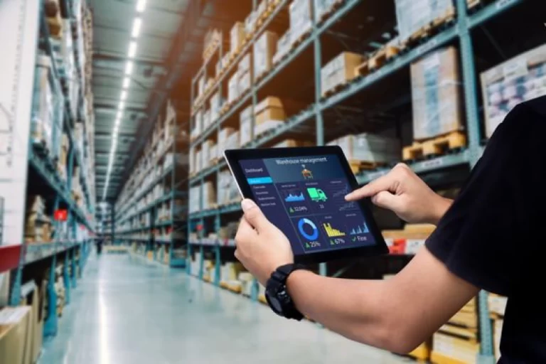 A person holding a tablet displaying a warehouse management system (WMS) in a large, organized warehouse with shelves stacked high with inventory, representing IT solutions for optimizing logistics operations.